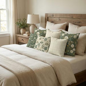 May include: A bed with a wooden headboard and a cream-colored comforter. Several decorative pillows in various patterns, including floral and plaid designs, are arranged on the bed. A bedside table with a lamp and a vase of greenery is visible.