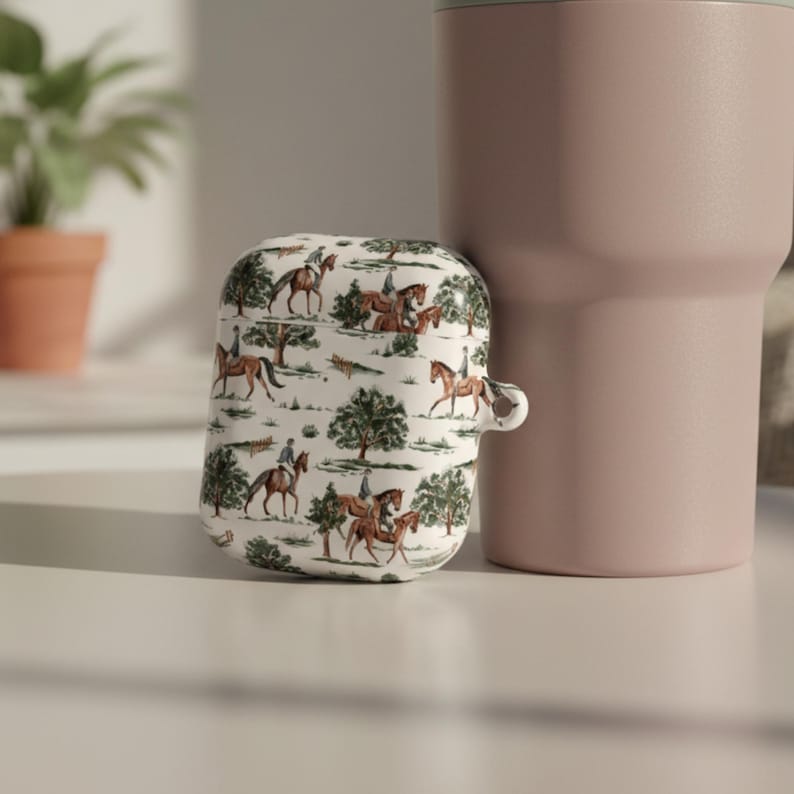 May include: White AirPod case with a repeating equestrian scene in brown and green. The case is next to a large pink tumbler. The design features riders on horseback, trees, and a landscape.