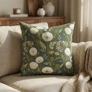 May include: A square decorative pillow with a dark green background and a floral pattern. The pattern features white dandelions, yellow flowers, and green leaves. The pillow is placed on a beige sofa.
