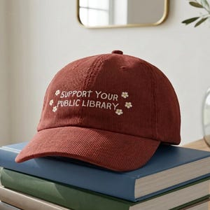 May include: A corduroy baseball cap in a deep red hue, embroidered with the phrase "SUPPORT YOUR PUBLIC LIBRARY" in white script, accented with small floral designs. The cap sits atop a stack of books.
