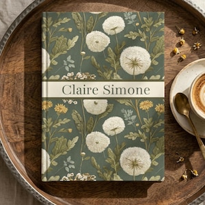 May include: A hardcover journal with a dark green background featuring a botanical pattern of white dandelion seed heads and small yellow flowers. A horizontal white band across the center displays the name Claire Simone in dark gray text.