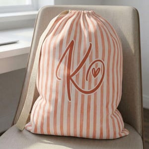 May include: A drawstring laundry bag featuring a vertical peach and white striped pattern. The front of the bag displays a large, dark orange cursive letter K with a small heart symbol integrated into the design. The bag has a thick, light-colored fabric strap.