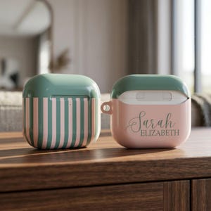 May include: Two protective cases for wireless earbuds. One case features vertical sage green and light pink stripes. The other case is solid light pink with a sage green lid and the name Sarah Elizabeth printed in a dark green cursive script font.