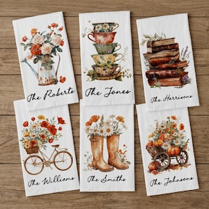 May include: Six white rectangular kitchen towels featuring watercolor illustrations. Designs include a floral watering can, stacked teacups, a stack of books, a bicycle with flowers, rain boots with flowers, and a wagon with pumpkins and flowers.