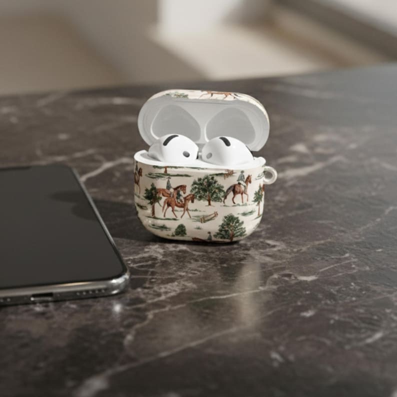 May include: An open, cream-colored AirPods case with a horse-themed design, containing white earbuds. The case features a repeating pattern of riders on horseback, trees, and foliage. A smartphone is visible in the background.