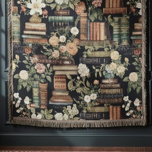May include: A rectangular woven blanket featuring a dark black background with a repeating pattern of stacked vintage books in various colours interspersed with clusters of white, pink, and orange flowers and green foliage, finished with fringed edges.