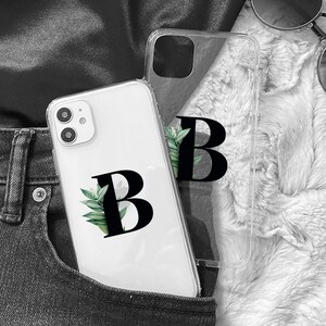 May include: A white phone case with a clear back and a black letter "B" with green leaves around it. The phone case is in a back pocket of a pair of black jeans.