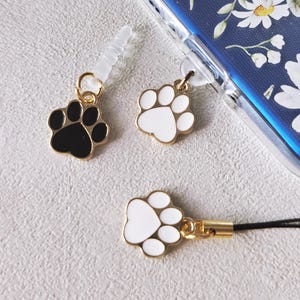 May include: A collection of paw print charms in black and white with gold-colored accents. One charm is attached to a black lanyard. A phone case with a floral design is also present.
