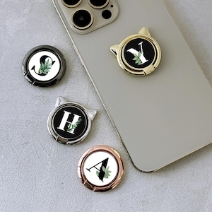 Op de afbeelding: Een verzameling telefoonhouders met initialen in goud, zilver en roségoud. Elke houder heeft een letter met een botanisch ontwerp. Eén houder is bevestigd aan een zilveren telefoon.