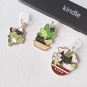 Könnte beinhalten: Drei dekorative Charms mit Katzenmotiven. Jeder Charm zeigt eine schwarze Katze, die aus Grün oder Blumen hervorlugt. Die Charms sind goldfarben mit schwarzen, grünen und rosa Emaille-Details. Im Hintergrund befindet sich ein Kindle-E-Reader.