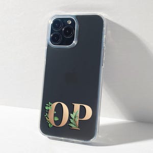 Pode incluir: Uma capa transparente para telefone com fundo preto e um monograma 'OP' dourado com folhas verdes em torno das letras.