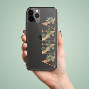 Könnte beinhalten: Transparente Handyhülle mit dem Namen "ANNA" in goldener Schrift, verziert mit grünen Blättern. Das Telefon wird in einer Hand vor einem hellgrünen Hintergrund gehalten. Das Telefon hat eine Dreifach-Kamera.