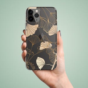 May include: Clear phone case with a black background and gold ginkgo leaf design. The case is held in a hand against a light green background. The leaves have detailed veins.