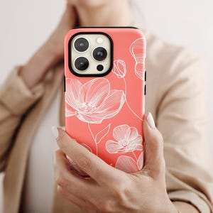 May include: Coral-colored phone case featuring a white floral design. The case has a black border and a camera cutout. The floral pattern includes detailed line drawings of flowers and stems. The phone case is held in front of a person.