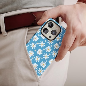 Puede incluir: Una funda de teléfono azul con un patrón de margaritas blancas es parcialmente visible en un bolsillo. La funda del teléfono la sostiene una persona que lleva un reloj marrón y un cinturón marrón. Las margaritas tienen centros amarillos.