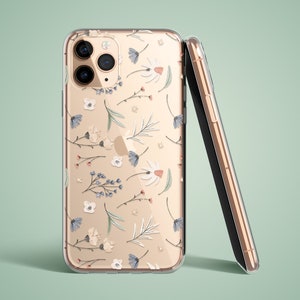 Puede incluir: Funda transparente para teléfono con un estampado floral de margaritas blancas, flores azules y hojas verdes. La funda está sobre un teléfono dorado, con un segundo teléfono apoyado. El fondo es verde claro.