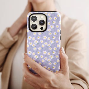 Può includere: Custodia per telefono con motivo a margherite. La custodia ha uno sfondo lilla chiaro con un motivo ripetuto di piccole margherite bianche con centri gialli e foglie verdi. La custodia del telefono è tenuta in mano da una persona.