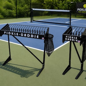 May include: Two black metal Pickle-Caddie ball storage racks with slatted tops and attached black mesh bags. The larger rack features the text PICKLE-CADDIE, while the smaller rack features the text PICKLE CADDIE, both with a small USA flag icon.