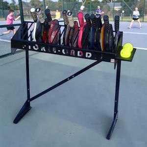 Puede incluir: Un estante de metal negro para palas de pickleball con múltiples palas de varios colores y diseños. El estante tiene un estante para pelotas de pickleball y el texto "PICKLE-GRID" es visible. Dos pelotas de pickleball amarillas están en el estante.