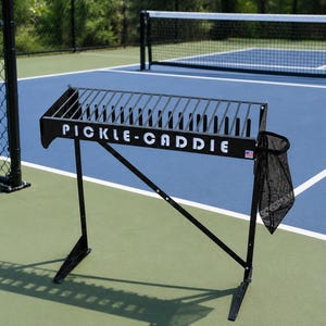 May include: Black metal pickleball caddie with the text "PICKLE-CADDIE" in white. The caddie has a rack for holding pickleball paddles and a mesh bag. The caddie is on a blue and green pickleball court.