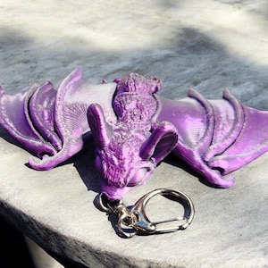 May include: A purple 3D printed bat keychain with a silver key ring. The bat is detailed with wings and a textured surface.