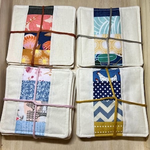 May include: A collection of four square fabric coasters. Each coaster features a white base with a central strip of colourful patterns. The patterns include floral, geometric, and illustrated designs. The coasters are bound together with twine.