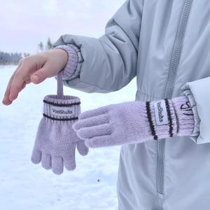 May include: A pair of lavender knitted gloves with black stripes and the text "VonShults" on the wrist. One glove is held by a loop, the other is on a person's hand. The background is a snowy landscape.