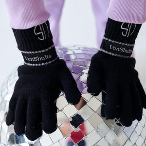May include: Black knit gloves with the text "VonShults" on the wrist. The gloves have a silver stripe and a stylized "S" logo. The gloves are shown against a mirrored disco ball.