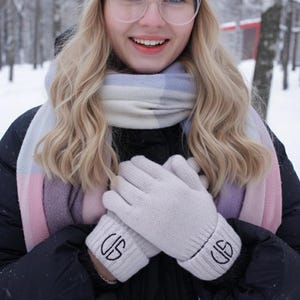 May include: A person wearing a gray knit hat, glasses, and a scarf with pink, blue, and white stripes. They are also wearing light gray gloves with black embroidered initials. The background is a snowy outdoor scene.