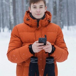 May include: A person wearing an orange puffer jacket with a hood, black gloves, and holding a smartphone. The gloves have a white stripe and text. The background is a snowy forest.