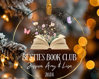 Peut inclure: Une boule de Noël en verre transparent avec un bord doré représente un livre ouvert avec des fleurs et des papillons qui émergent des pages. Le texte "Besties Book Club Jessica, Amy & Lisa 2024" est imprimé sur la boule.