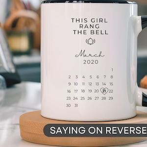 May include: White ceramic mug with a black handle. The mug has black text that reads "This girl rang the bell" and a calendar for March 2020 with the 21st circled. The mug is sitting on a wooden coaster with the text "Saying on reverse side".