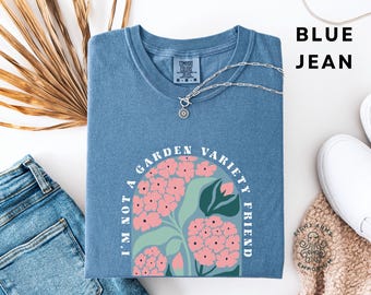 May include: Blue jean coloured t-shirt with the text "I'm not a garden variety friend" and a floral design in pink and green. A silver necklace with a pendant is on the shirt. The shirt is folded and placed on a white surface.