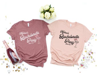 May include: Two short-sleeved t-shirts, one mauve and one light pink, with the text "Melissa's Bachelorette Party July 2025" in a script font. A champagne bottle and confetti are scattered around the shirts. A bouquet of white flowers is in the background.