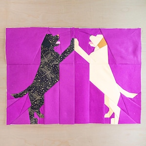 May include: A fabric art piece featuring two stylized dogs in a high-five pose. One dog is black with a gold dragonfly pattern, the other is cream-colored. The background is a vibrant purple.