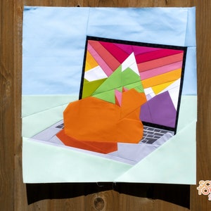 Puede incluir: Una colcha de retazos colorida que representa un portátil con una escena de montaña en la pantalla. La colcha está hecha con varios tonos de azul, verde, naranja, morado y rosa. La colcha está firmada "Scrap Tower Studio" en la esquina inferior derecha.