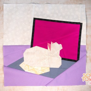 May include: A fabric art piece with a geometric design. It features a white origami-style cat on a grey surface, set against a bright pink backdrop with a black border. The design incorporates lavender, yellow, and white patterned fabrics.