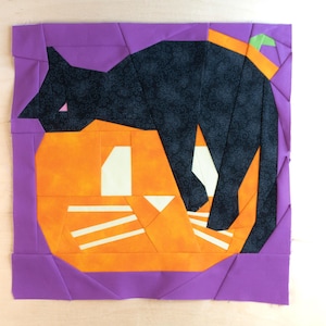 Peut inclure: Un bloc de tissu violet avec un chat noir enroulé sur un potiron orange. Le chat a un nez rose et le potiron a des rayures blanches.