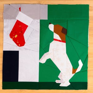 May include: A patchwork Christmas-themed block featuring a red stocking with gold accents and a dog with brown and white markings. The dog is wearing a red collar and is set against a green background.