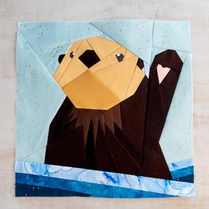 May include: A brown and tan sea otter with a pink nose and a blue and white background. The otter is made of fabric and is a patchwork design.