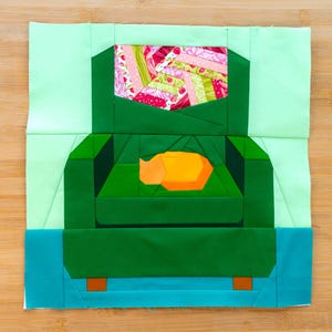 May include: A colourful quilt block showing a green armchair with an orange cat inside. The chair is set against a light green background, with a patterned pillow. The block is on a wooden surface.