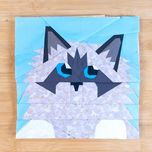 May include: A square fabric art piece featuring a stylized cat face. The cat has blue eyes, gray ears, and a white and patterned face. The background is light blue. The artwork is placed on a wooden surface.