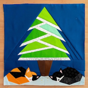May include: A fabric artwork depicting a green Christmas tree with white detailing against a blue backdrop. Two stylised cats, one orange and black, the other black, are resting beneath the tree. The artwork is a geometric design.