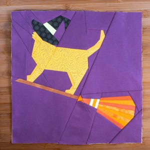 Puede incluir: Un diseño de patchwork geométrico con un gato amarillo con un sombrero de bruja, montado en una escoba sobre un fondo púrpura. La escoba tiene rayas naranjas y amarillas. El gato está hecho de tela amarilla con un pequeño estampado floral.