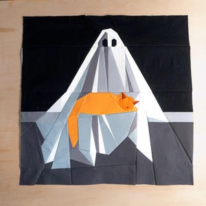 Könnte beinhalten: Ein Textilkunstwerk mit einem geometrischen Geist und einer schlafenden orangefarbenen Katze. Der Geist ist weiß mit schwarzen Augenlöchern, und die Katze liegt in seinen Falten. Der Hintergrund ist schwarz und grau.