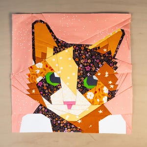 Può includere: Un blocco di tessuto quadrato con un design geometrico di gatto. Il volto del gatto è composto da vari modelli di tessuto, tra cui floreali, pois e colori uniti in arancione, giallo, nero e bianco. Il gatto ha occhi verdi e un naso rosa.