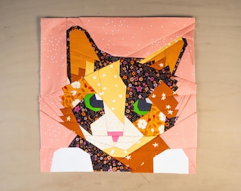 May include: A square fabric block featuring a geometric cat design. The cat's face is composed of various fabric patterns, including floral, polka dots, and solid colors in orange, yellow, black, and white. The cat has green eyes and a pink nose.