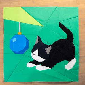 Op de afbeelding: Een patchwork ontwerp met een zwart-witte kat die naar een blauwe bal grijpt. De achtergrond is groen, met een lichtgroene driehoek. De kat heeft witte poten en een grijs oor.