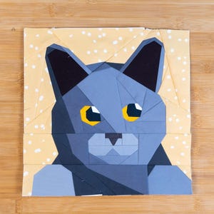 May include: A geometric cat design on a square fabric block. The cat is primarily blue with yellow eyes and black ears. The background is a light yellow fabric with white polka dots. The design is likely for a quilt or other fabric craft.