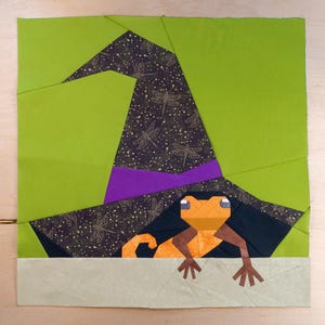 May include: A fabric quilt block featuring a pointed witch hat in black fabric with a gold dragonfly pattern and a purple band. An orange lizard with brown limbs is positioned beneath the hat against a bright lime green background.